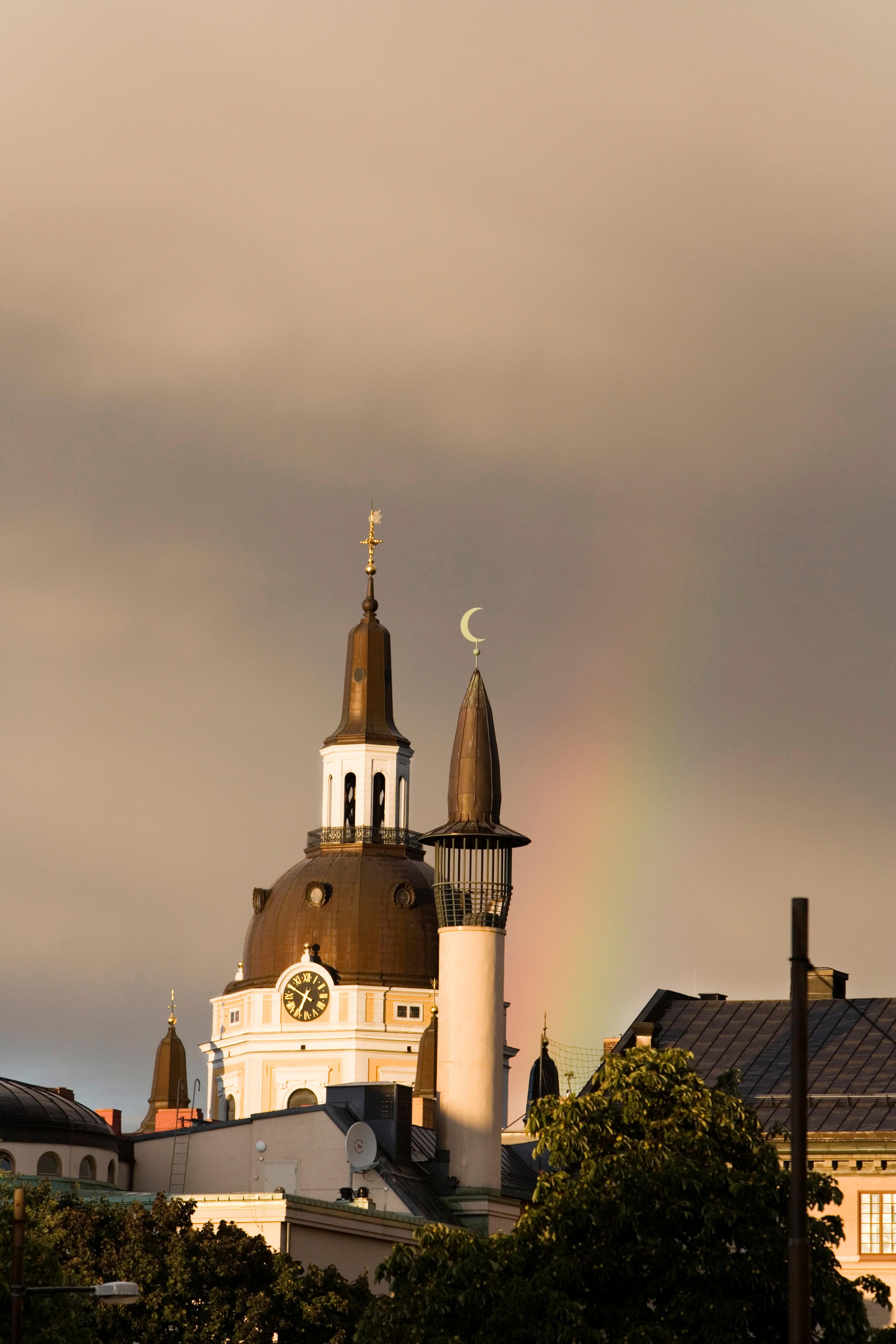 برجين لكنيسة ومسجد تحت سماء داكنة مع قوس قزح في الخلفية.